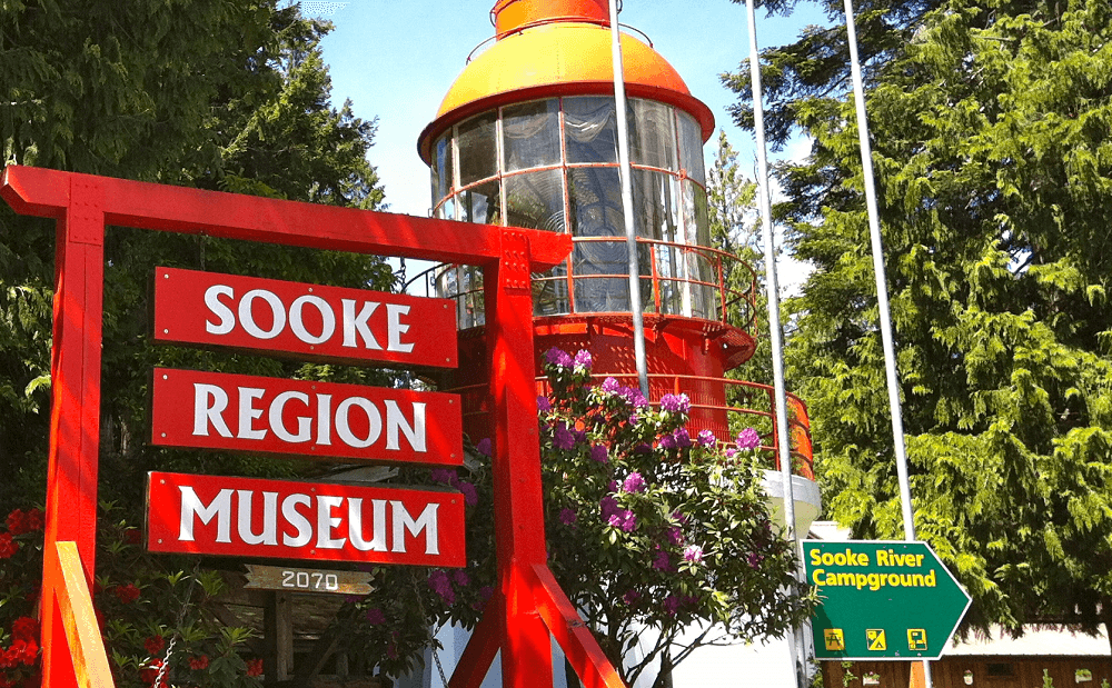 The Sooke Region Museum red sign with the lighthouse and flag poles in the background.